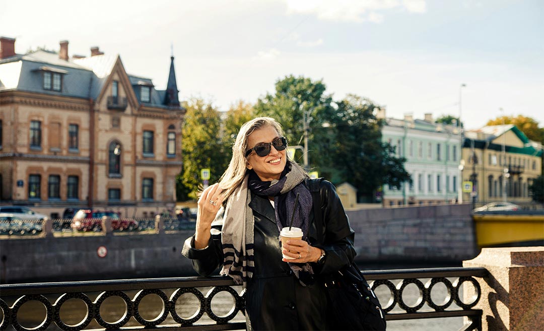 A confident woman standing on a historic stone bridge over a European river, smiling and holding a drink during a solo journey.