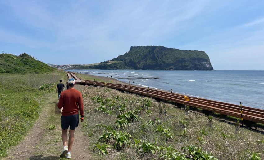 Sofia's travel companion walking along a scenic dirt path by the sea, with a majestic volcanic crater rising on the horizon ahead under a soft sky.