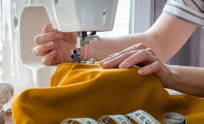 Close-up of a woman’s hands carefully guiding fabric through a home electric sewing machine, highlighting self-reliance and learning practical life skills during a career break.