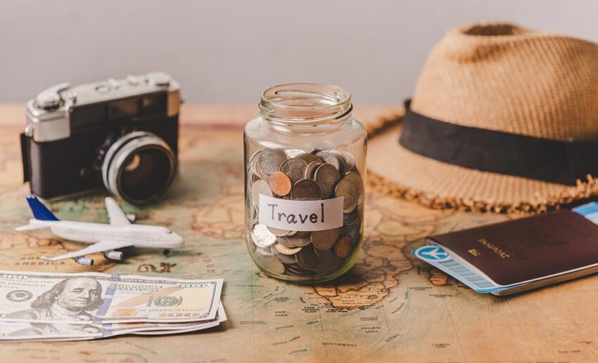 A clear glass savings jar filled with coins and rolled banknotes resting on a world map next to a passport, illustrating how to build a travel savings fund.
