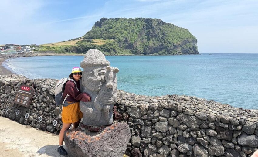 Sofia smiling and leaning companionably next to a traditional Dol Hareubang volcanic stone grandfather statue on Jeju Island.