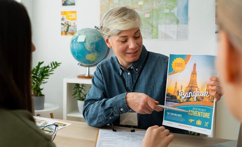 Front view of a female travel agent smiling and holding up a Bangkok adventure holiday pamphlet at a busy tourism exhibition or travel expo.