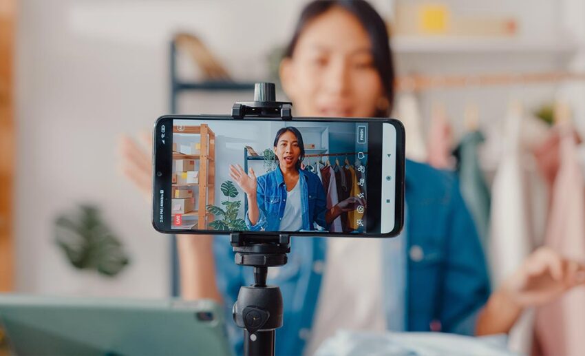 A female small business entrepreneur live streaming clothing items on a smartphone, representing creative independence and testing out a side business idea.