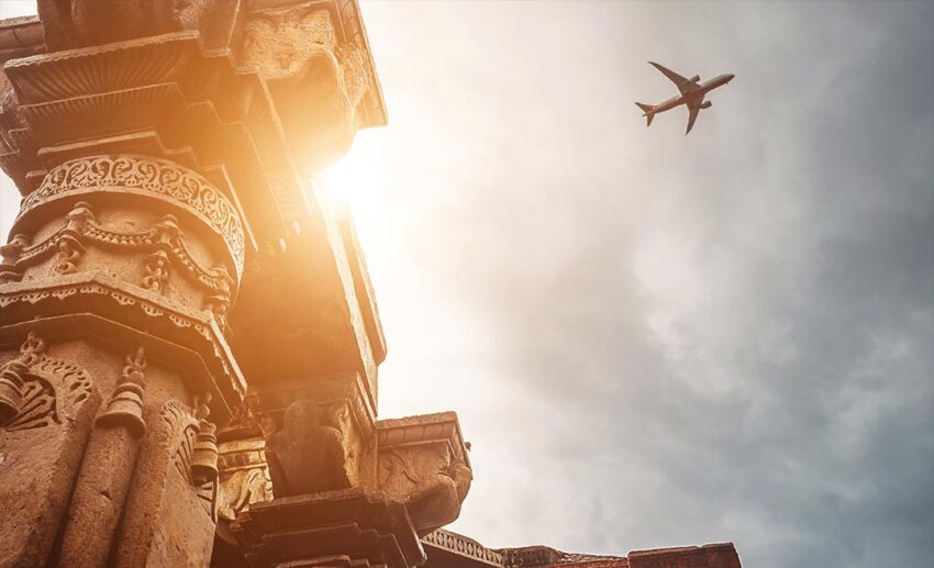 An airplane flying high over the historic stone pillars of the Qutb Minar complex against a clear blue sky in Delhi, India.