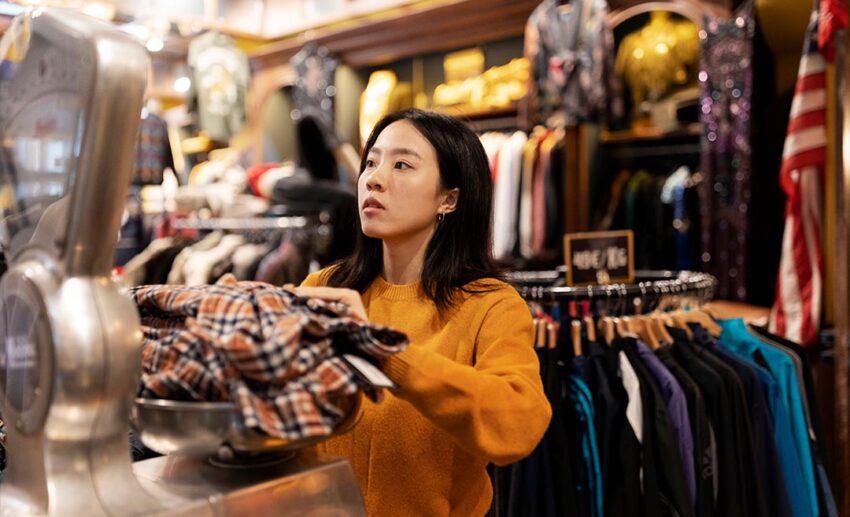 Side view of a woman browsing through racks of pre-loved clothing at a local thrift store, looking for affordable second-hand winter wear and travel gear.