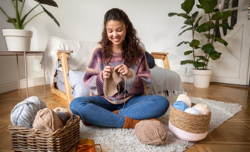 Full shot of a smiling woman sitting comfortably at home and knitting a colourful garment, exploring creative side hustles to earn extra money for a holiday fund.