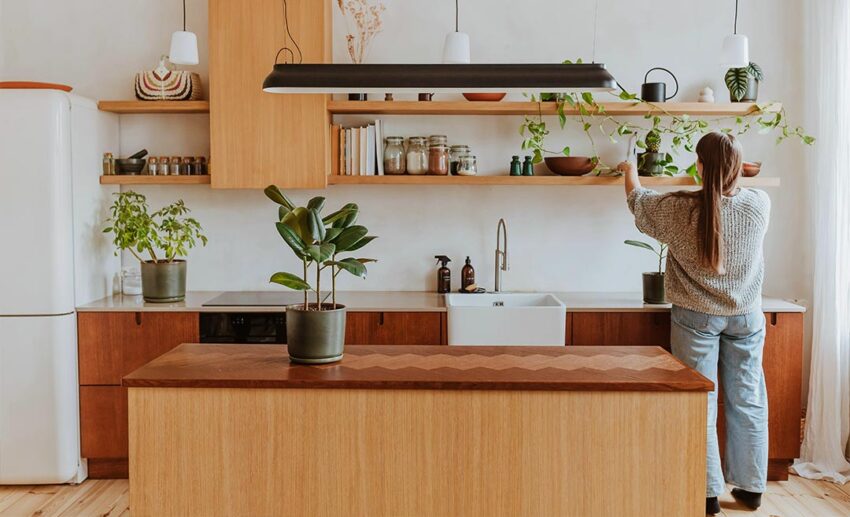 A woman standing in a bright, modern, and newly renovated kitchen while carefully placing a potted plant on open wooden shelving above clean wooden countertops.