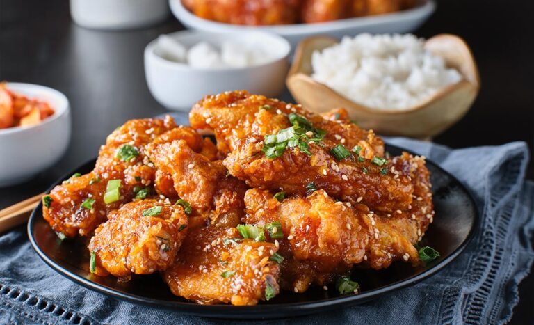A close-up shot of crispy fried Korean chicken wings glazed in a glossy galbi sauce, served on a table with small banchan side dishes including pickled radish, kimchi, and a bowl of steamed white rice.