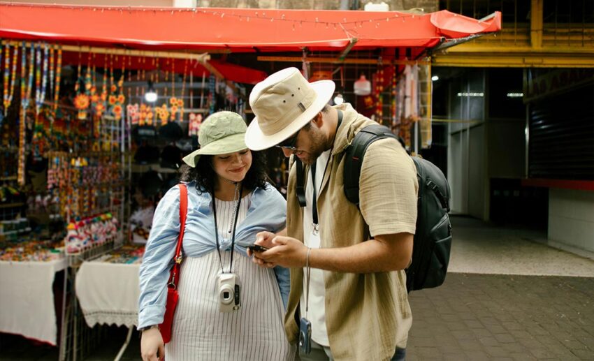 A young couple standing in a vibrant outdoor market together, looking down at a mobile phone screen to track travel spending and redeem local cashback rewards.