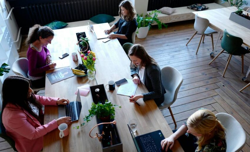A group of women gathered around a wooden table sharing laptops, papers, and travel planning brochures