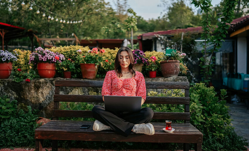 A minimalist female traveller sitting with her legs tucked on a bench, working on a laptop with minimal luggage.