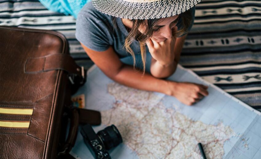 A cheerful woman sitting comfortably at home, smiling as she traces a route on a large paper travel guide map, embodying a happy wanderlust and independent lifestyle.