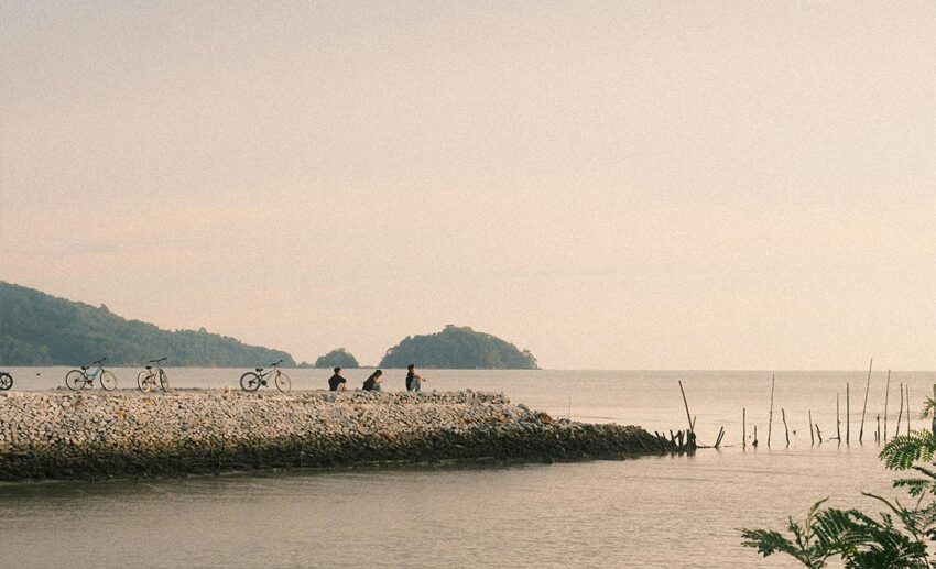 Three cyclists resting on a rocky coastal bay at dusk under an orange sky with parked bicycles nearby