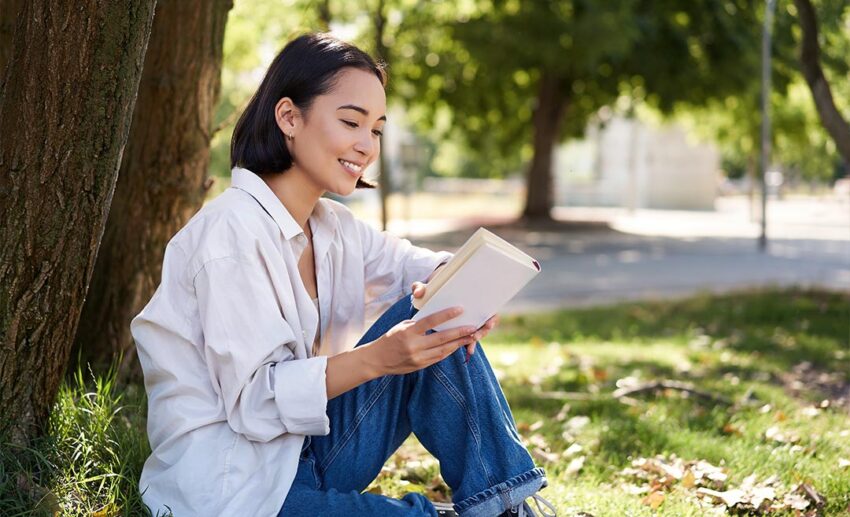 A young woman sitting peacefully under a tree in a green park reading a book, demonstrating the grounding effect of spending time in nature.