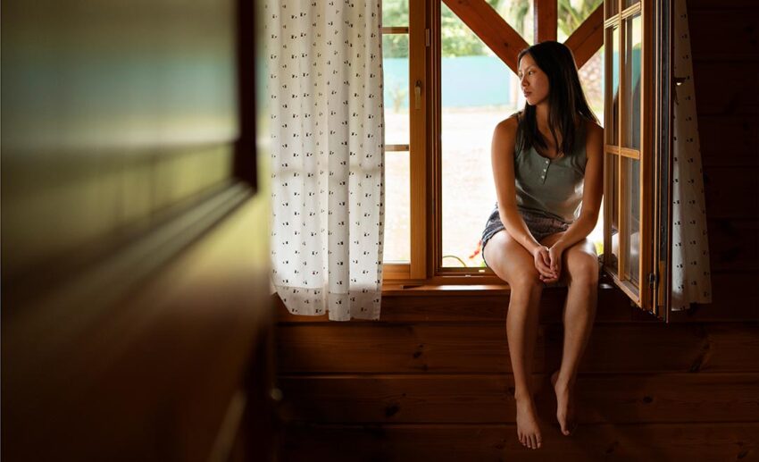 woman sitting quietly on a windowsill looking inwards, portraying the fatigue and hesitation that often accompany grief before taking a trip.
