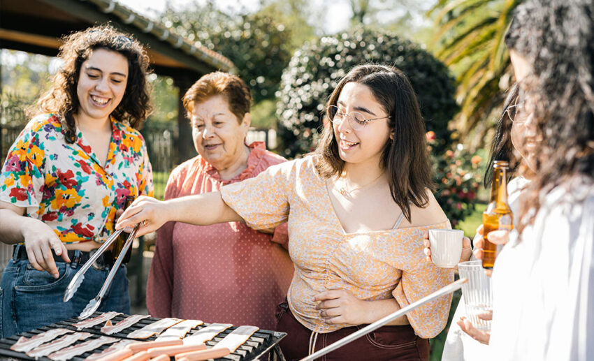 A group of women gathering around an outdoor barbecue grill during a local social event, sharing food and drinks.