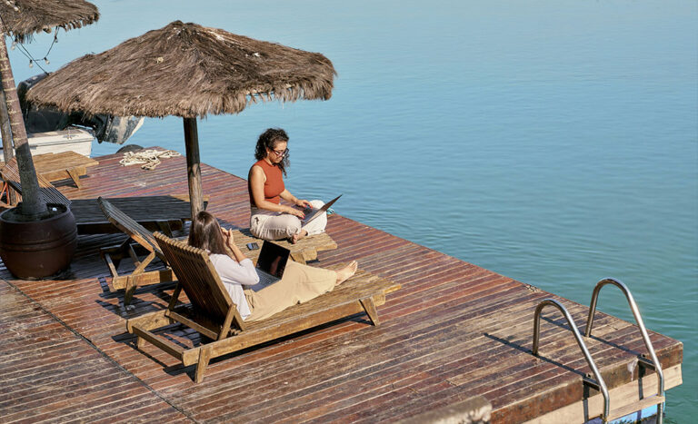 Two digital nomad women working remotely on laptops while sitting on wooden lounge chairs on an outdoor deck overlooking open water.