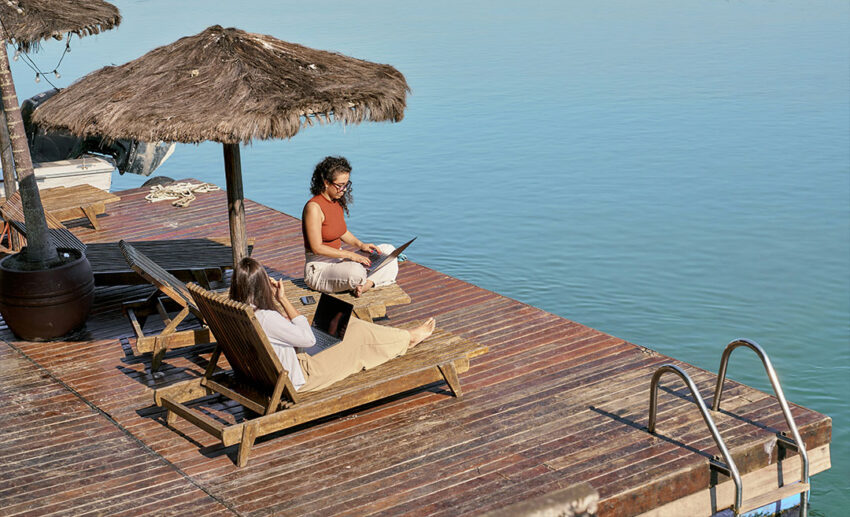 Two digital nomad women working remotely on laptops while sitting on wooden lounge chairs on an outdoor deck overlooking open water.