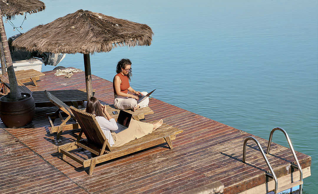 Two digital nomad women working remotely on laptops while sitting on wooden lounge chairs on an outdoor deck overlooking open water.