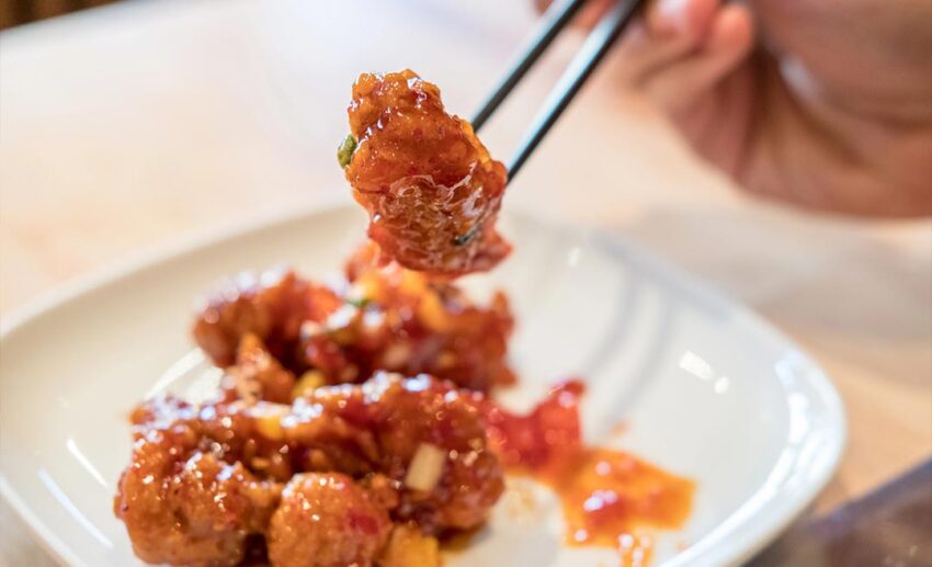 A close-up view of a person using metal chopsticks to lift a piece of golden, crispy seasoned Korean fried chicken from a serving dish.