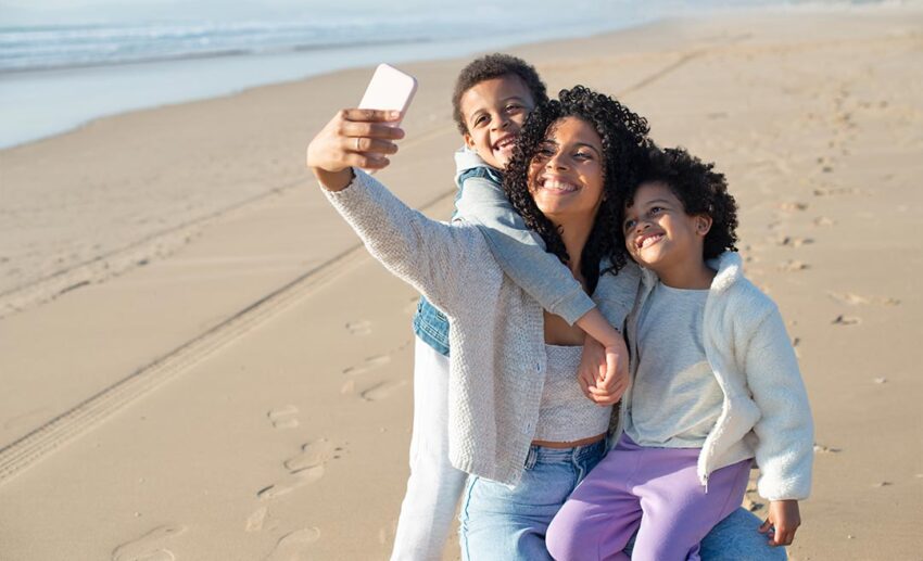 A mother smiling happily while taking a casual beach selfie with her children, capturing meaningful family moments and making gentle new travel memories.