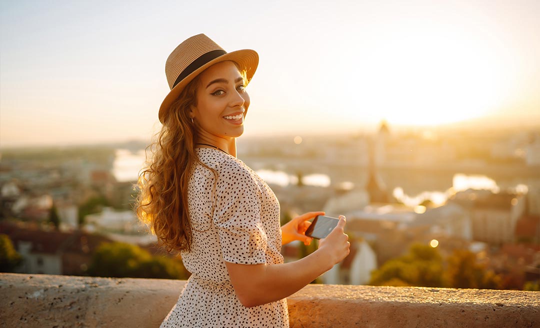 A female tourist looking over her shoulder at the camera while preparing to take a photograph of a European city landscape at dawn, representing independent travel planning.