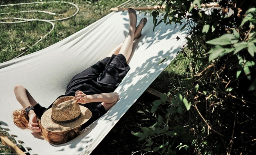 A woman resting peacefully in an outdoor hammock with a sun hat resting over her face, representing low-pressure travel and permitting oneself to rest.