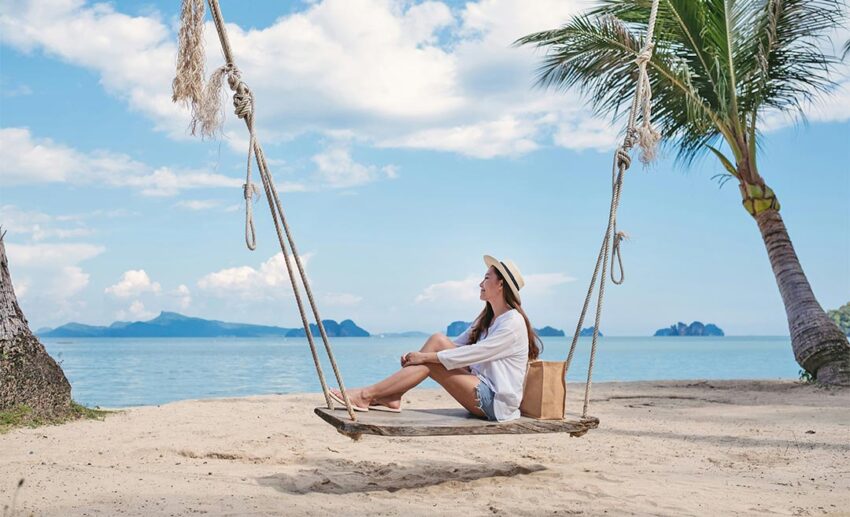 A contemplative young Asian woman sitting on a wooden swing by the sea beach, considering an adult gap year.