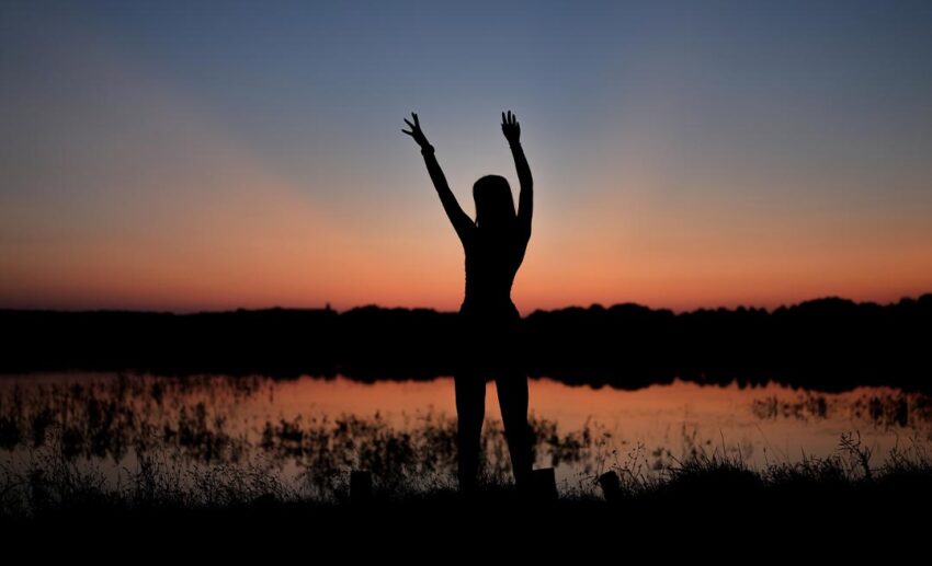 Silhouette of a woman standing with arms raised by a calm lake at sunset, reflecting on her emotional experience during an extended stay abroad.