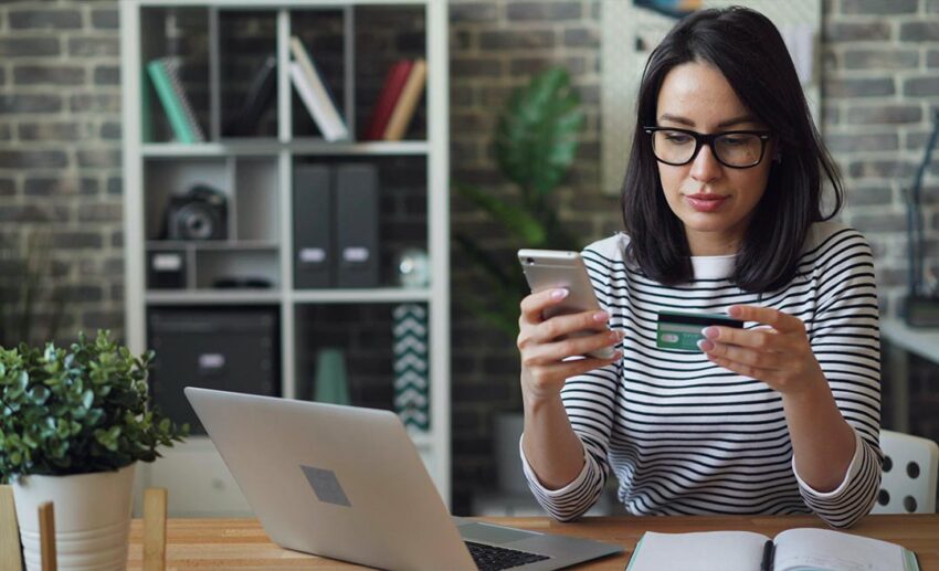 A woman sitting at a table checking her mobile phone screen while holding a credit card to manage expenses and budget her monthly cost of living.