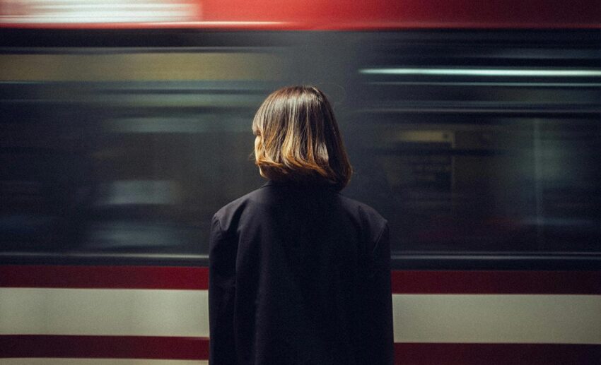 A woman standing patiently on a public transport station platform as a passenger train speeds past, illustrating a daily commute routine.