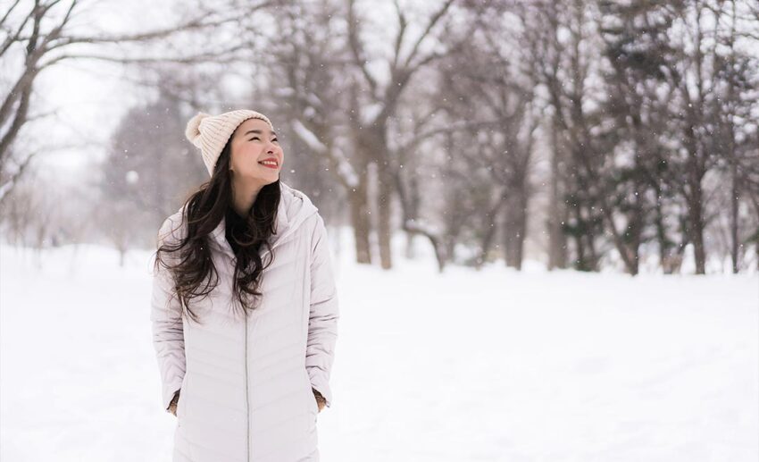 A smiling young Asian woman wrapped up in warm winter clothes enjoying a snowy travel landscape, showing how choosing a favourite season enhances travel comfort.
