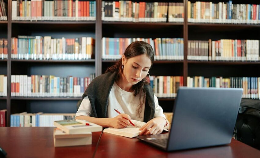 A woman studying independently at a library wooden table with a laptop, open textbooks, and a notebook, focusing on upskilling during a mid-career sabbatical.