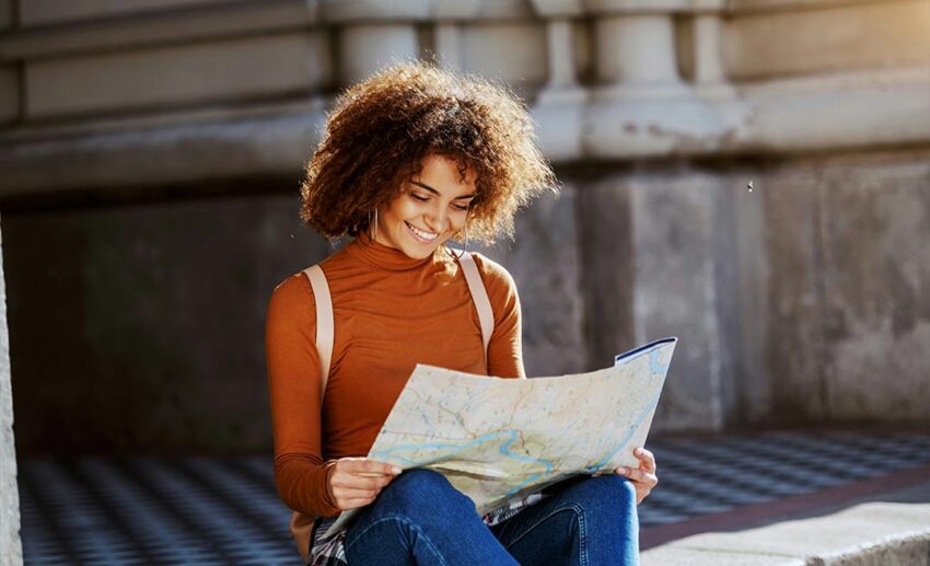 Mixed-race female tourist with curly hair sitting on a city street curb, studying a paper map, and looking for directions