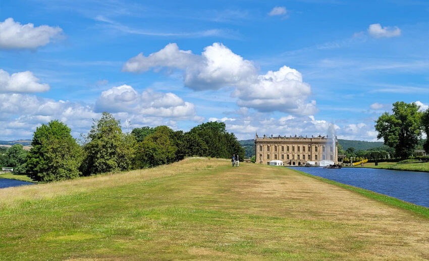 Distant view of Chatsworth House across a calm lake, surrounded by green hills in the Derbyshire Peak District