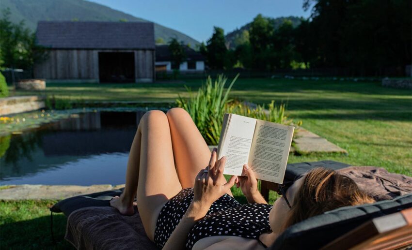 Woman in a polka dot dress relaxing on a deck chair while reading a book beside a tranquil pond