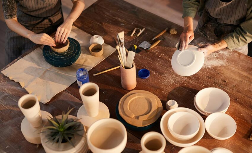 Close-up of female artisans wearing aprons and shaping clay ceramics at a wooden workshop table.