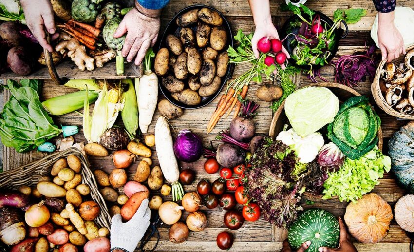 High-angle shot of four pairs of hands selecting fresh organic vegetables laid out on a rustic wooden market table