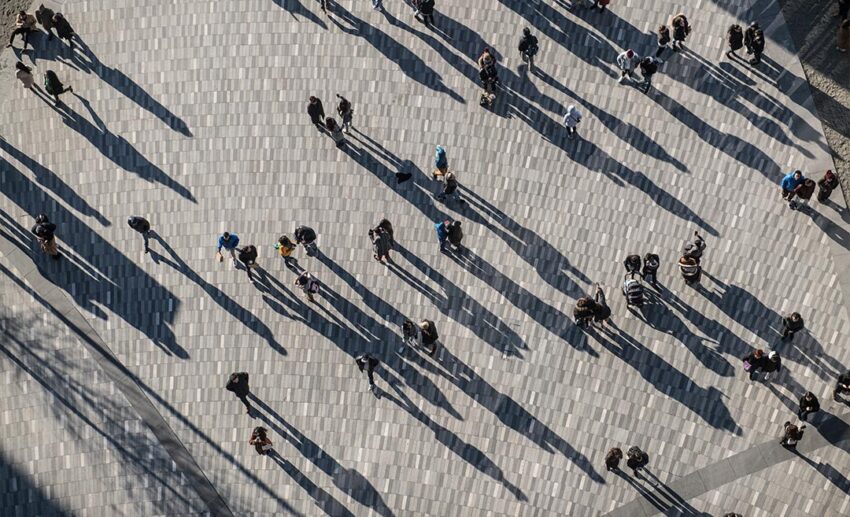Top-down aerial view of pedestrians walking across a crowded city square casting long shadows in the sunlight