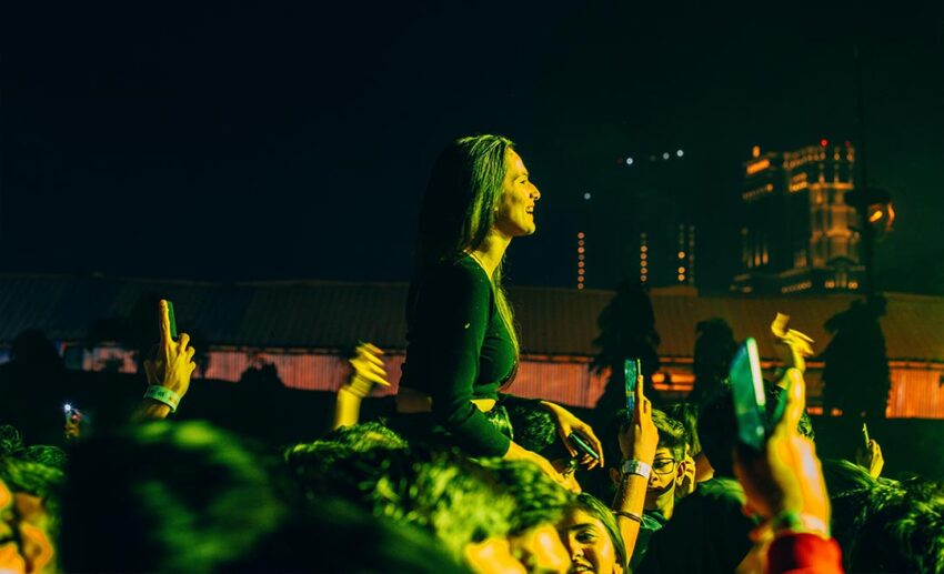 Woman sitting on a friend’s shoulders celebrating among a crowd at a night music event in Kolkata, India.