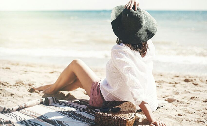 Woman wearing a sun hat relaxing on the beach next to a straw bag near ocean waves.