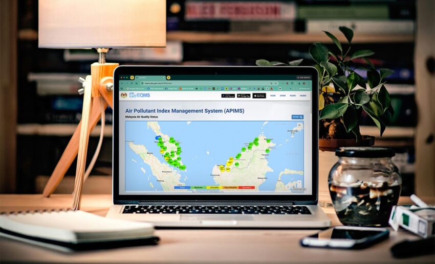 A MacBook Pro resting on a brown wooden table indoors, displaying the Department of Environment's official Air Pollutant Index Management System (APIMS) website on its browser screen