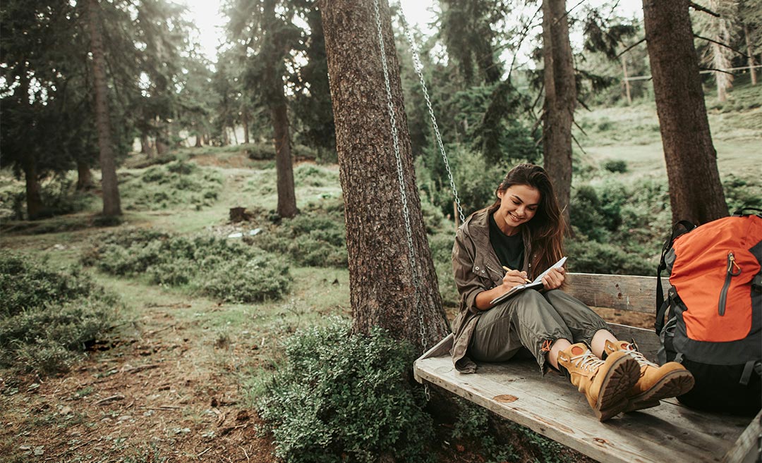 Woman writing in a paper notebook, smiling, and sitting on a wooden bench surrounded by trees in a quiet forest
