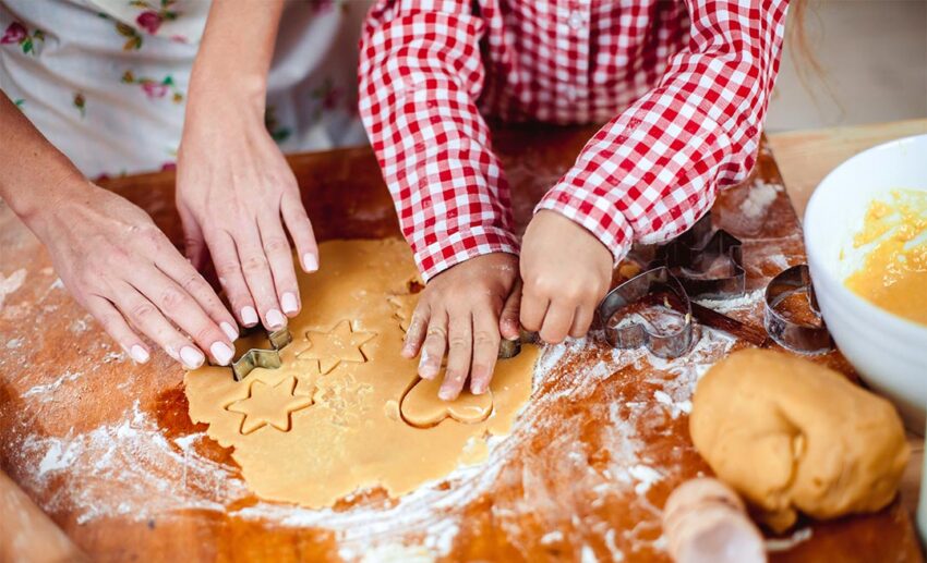 Close-up of a mother and daughter's hands shaping cookie dough together in a warm festive kitchen near a Christmas tree.