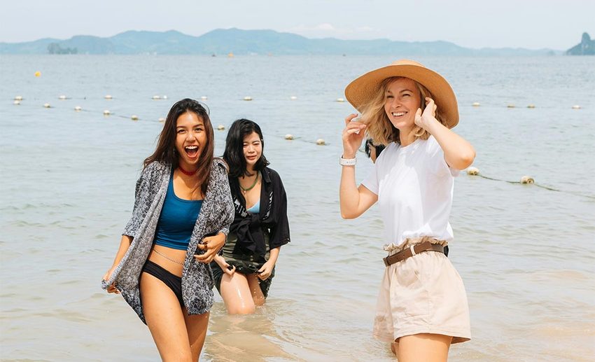 Three female friends walking arm in arm along shallow tropical sea waters, laughing together on a sunny beach.