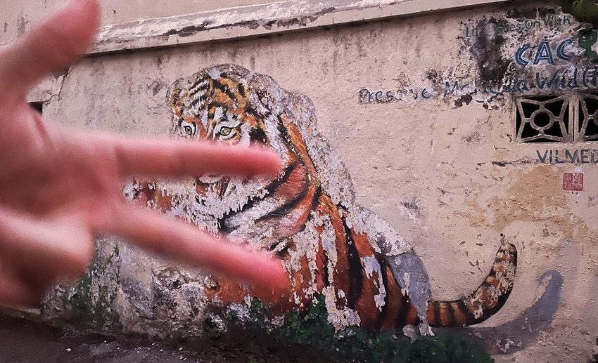 Hand held up in a playful gesture in front of a weathered wall featuring faded, vintage street art in Ipoh Old Town.