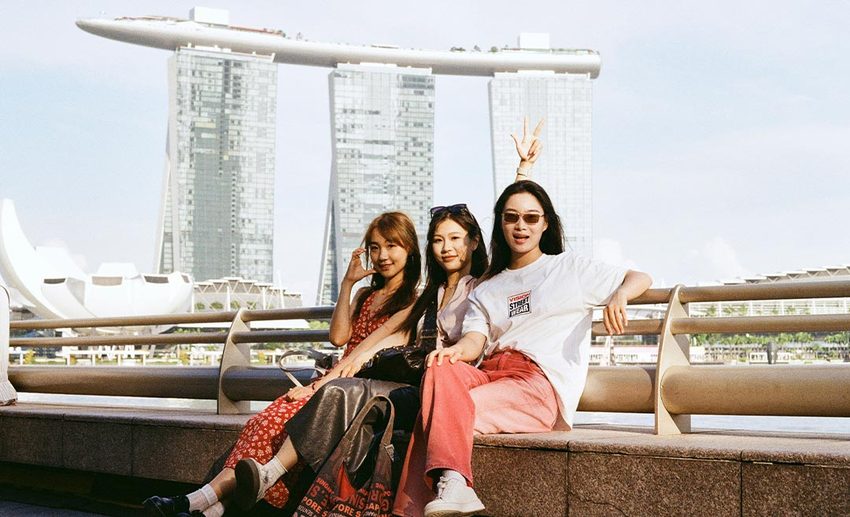 Three female travel friends sitting outdoors along the waterfront, posing with the iconic Marina Bay Sands skyline in Singapore.