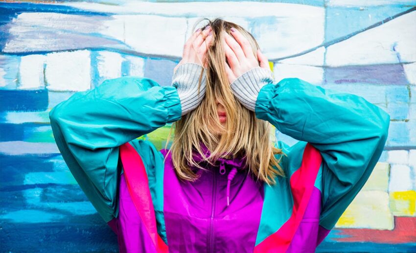 A young woman wearing a retro 1990s style jacket shyly covering her face with her hands and hair.