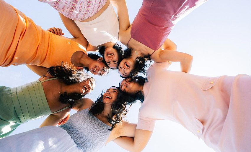 A group of women standing in a joyful circle with their arms around each other's shoulders, celebrating new friendships.