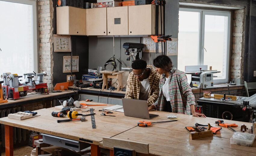 Group of participants collaborating on woodworking projects at a workshop workbench using hand tools, timber, and a laptop.